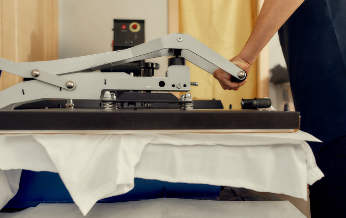 A person’s arms pushing down on heat press handle while the machine clamps down on a white t-shirt. 