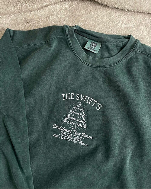 A long-sleeved C6014 Comfort Colors tee from ShirtSpace in the dark green color “Blue Spruce” and embroidered with “The Swift’s Christmas Tree Farm – Cut and Carry – Pine – Spruce – Fur -–Cedar” and a Christmas tree illustration, decorated by Hannah Collective. 