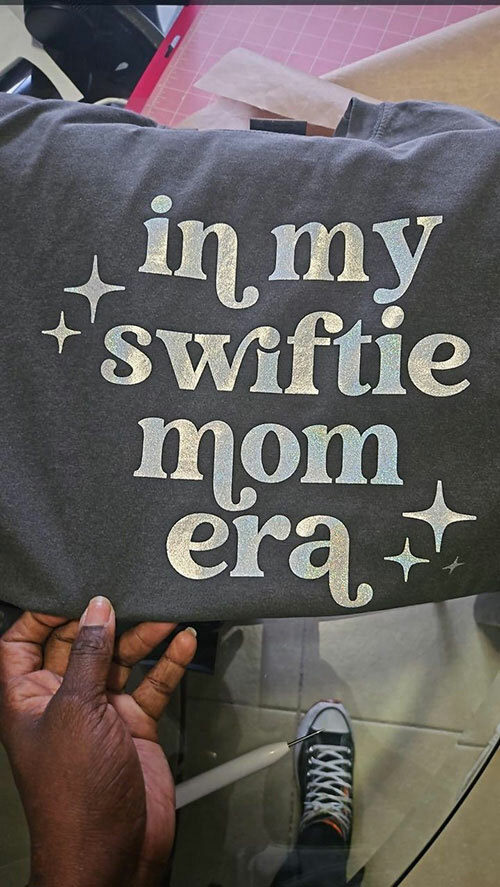 Dark gray t-shirt from ShirtSpace with a silver metallic print that reads: in my Swiftie mom era, decorated by trayseamakesthings.