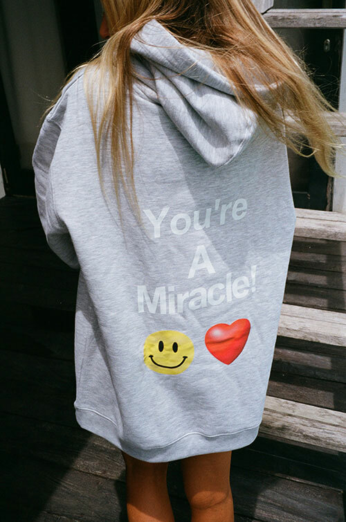 Girl with long blonde hair wearing an oversized ash gray colored sweatshirt that says “You’re a Miracle” with smiley face and heart emojis printed on the back.