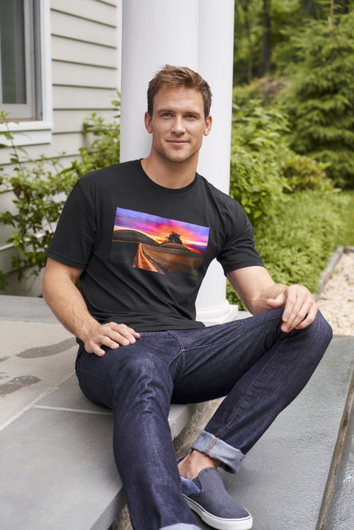 Man sitting on porch steps, wearing a black Gildan 64EZ0 t-shirt with a vibrant sunset print, and jeans.