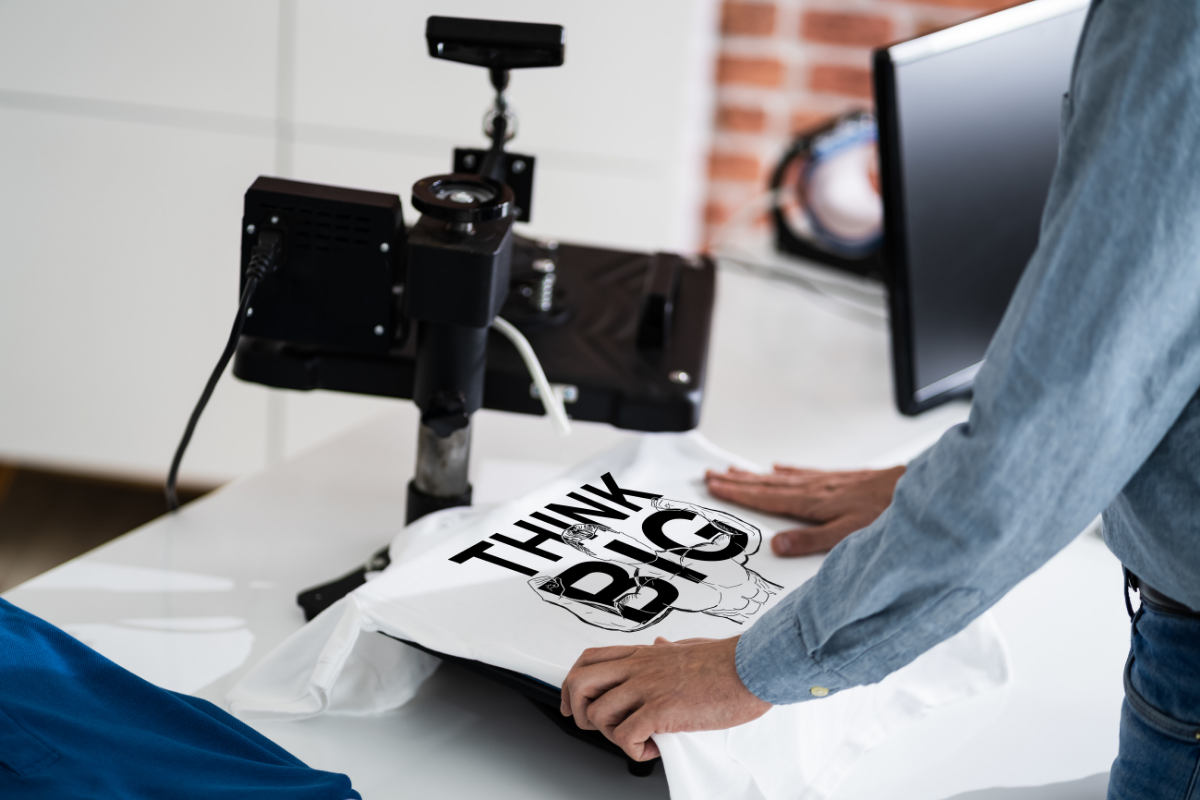 Person printing a t-shirt design that says "Think Big" in black writing on a white t-shirt from ShirtSpace.