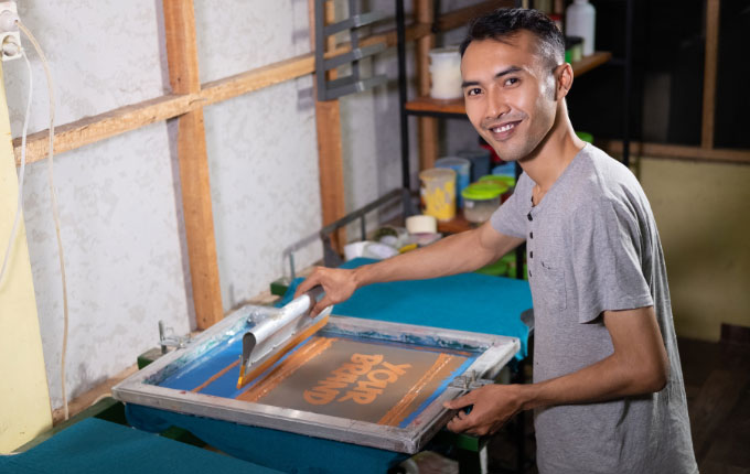 Man screen printing on t-shirts in an unfinished garage.