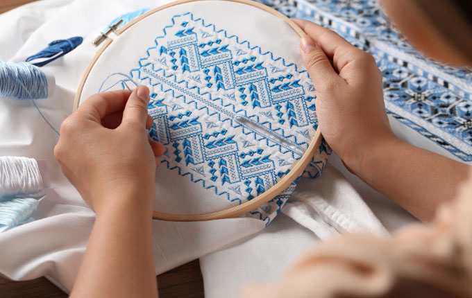 Person hand embroidering a design on white fabric with blue thread, using an embroidery hoop.