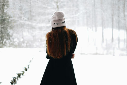 A woman standing in a snowy landscape while wearing a white embroidered beanie with cartoon-like eyelashes stitched onto the cuff. 