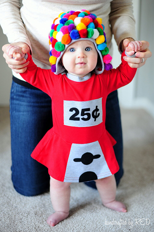 An infant wearing a red outfit that has been customized to look like a gumball machine, and a beanie covered in pom-poms, in order to look like gumballs. 