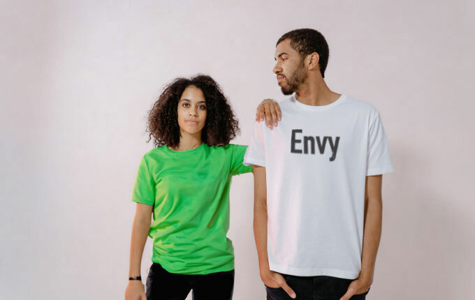 A woman in a green t–shirt, standing with her arm resting on the shoulder of a man who is wearing a white t-shirt that reads: “Envy”, to create the couple’s Halloween costume, “Green with Envy”.