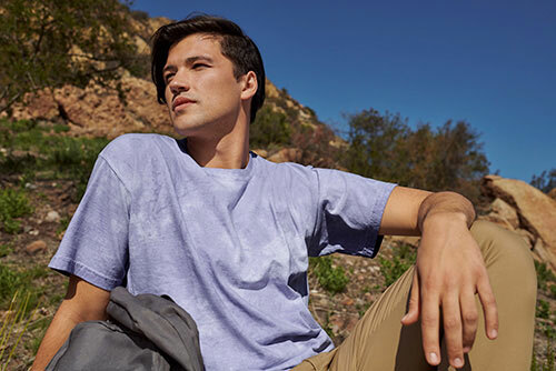 Man sitting on a rocky hillside, wearing the Comfort Colors 1745 Adult Heavyweight Color Blast T-Shirt in “amethyst” and a pair of khaki pants.