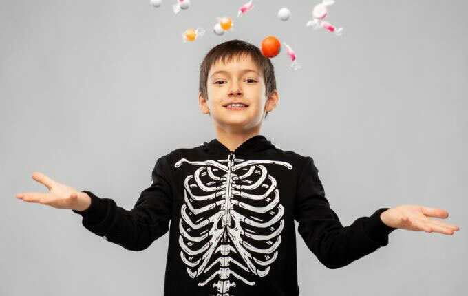 A boy wearing a black full-zip hoodie with a skeleton print on the front, throwing up candy into the air from trick-or-treating.