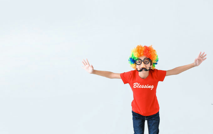 A child wearing a rainbow clown wig and glasses with a fake nose and mustache. She is also wearing a red-shirt that reads, “Blessing”, to finish off her “Blessing in Disguise” costume. 