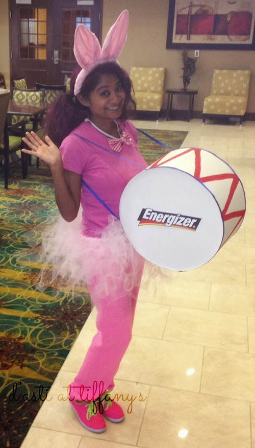 A woman wearing a pink t-shirt, sweatpants, tulle tutu skirt, and bunny ears while holding a drum with the Energizer logo – dresses as the Energizer Bunny for Halloween. 