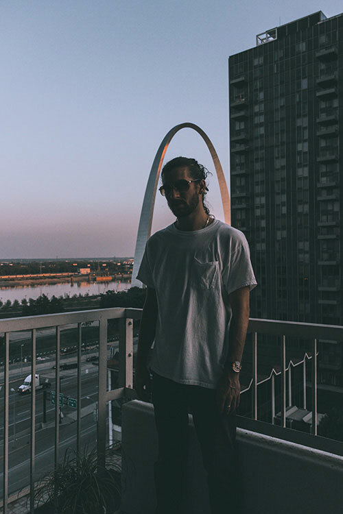 A man on a balcony with St. Louis arch in the background wearing a white pocket tee, styled with gold jewelry, dark pants and sunglasses.