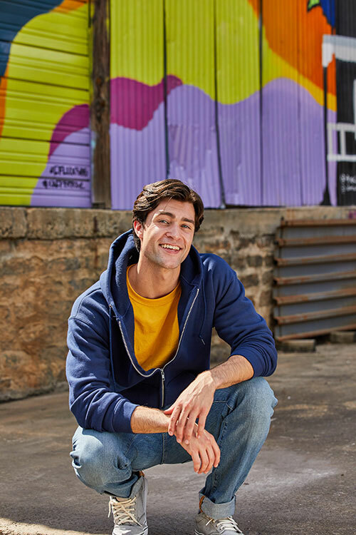 Man with his hands in his pockets wearing a mustard yellow t-shirt with a zip-up navy blue Fruit of the Loom sweatshirt. 