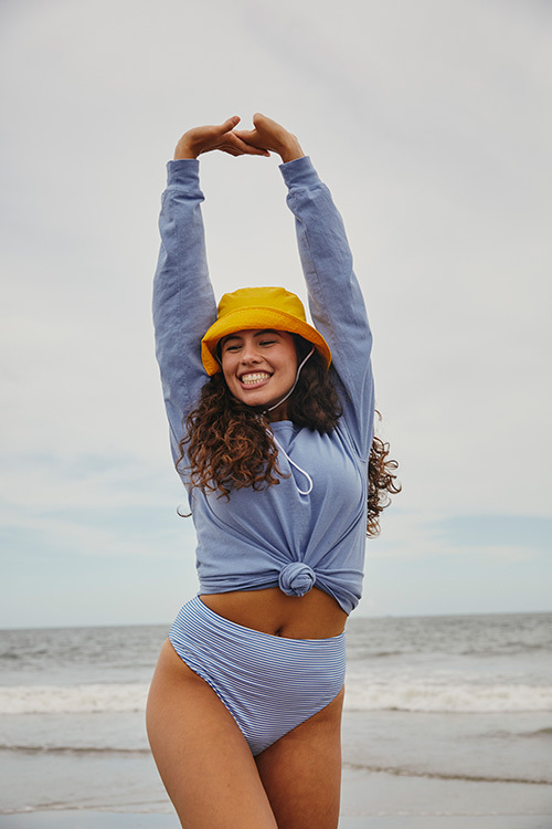 Girl sitting on a blanket on the beach wearing a long-sleeved ComfortWash by Hanes t-shirt in the color “saltwater”, paired with a bright gold bucket hat. 