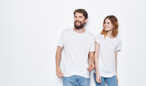 Man and woman standing in front of white background, wearing white t-shirts and blue jeans. 