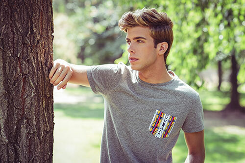 A young man wearing a heather gray pocket tee, with a colorful tribal printed pocket. 