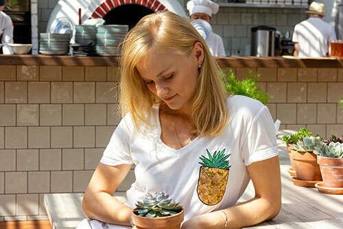 A woman holding a small potted succulent plant while wearing a white v-neck t-shirt that has a faux pocket, made to look like a potted plant. 