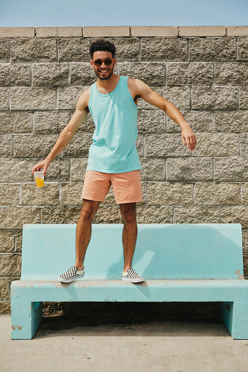 Man standing on a bench wearing a light blue GDH300 tank top from ComfortWash by Hanes, paired with peach-colored shorts. 