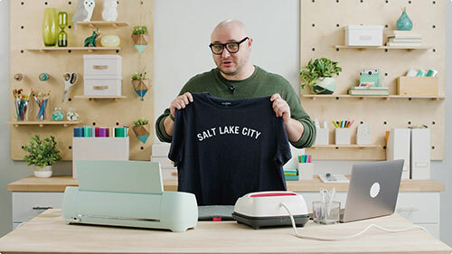 Person holding up a Salt Lake City t-shirt a craft studio with a laptop, Cricut vinyl cutter and heat press.