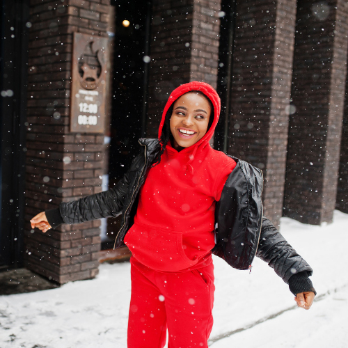 Black woman in a red hoodie and sweatpants and black jacket smiling in the falling snow 