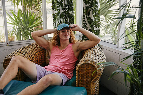 Man wearing a ComfortWash by Hanes GDH300 tank top in the color “Crimson Fall” paired with violet shorts while relaxing in a wicker chair.