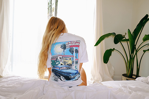 A woman showing off the back of her t-shirt, printed with a vintage picture of In-N-Out burger and a classic car. 