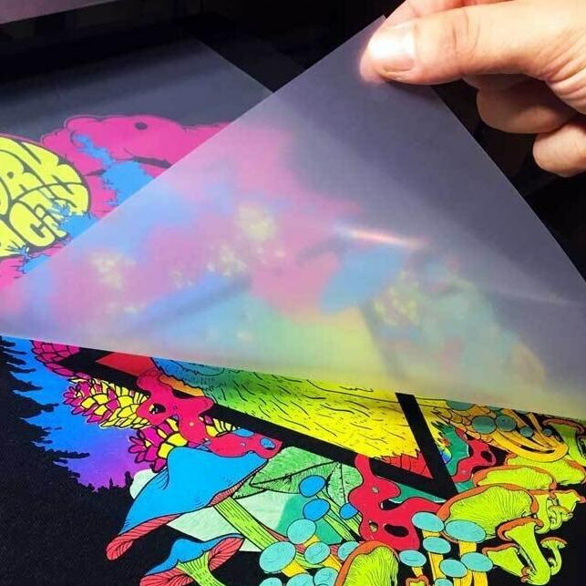 A hand peeling away the film from a DTF transfer, revealing a colorful, psychedelic print on top of black material.